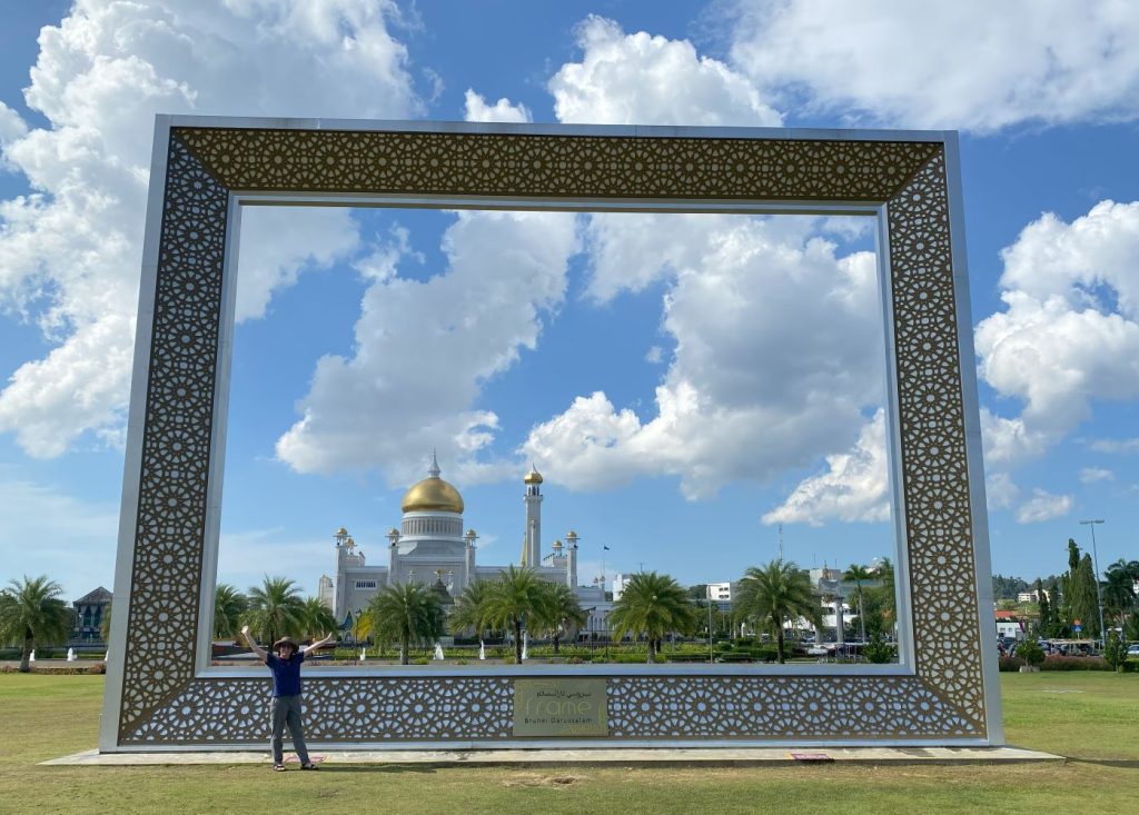 Me standing in front of a large frame-shaped sculpture with the Omar Ali Saifuddien Mosque in the background