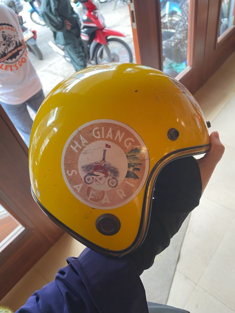 A yellow motorcycle helmet with a "Ha Giang Safari Hostel" sticker on it.