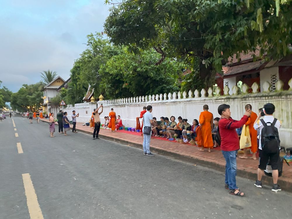 A line of petitioners sitting with offerings while monks in bright orange robes walk down the sidewalk. A handful of tourists with cameras stand close to take photos.