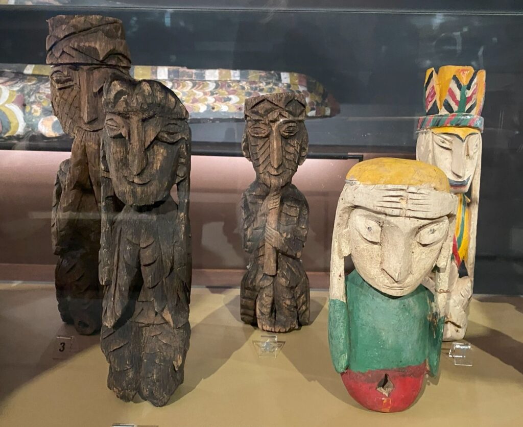 Five wooden figures carved in an outsider art style. Two are painted in primary colors and the other three are a darker stained wood color.