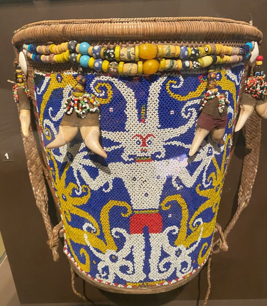 A large basket with a beaded design of a person and a beaded rim with animal teeth hanging off.