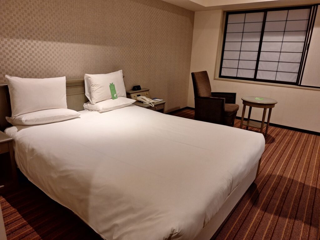 A large double bed in a hotel room with an armchair and a small side table in the back.