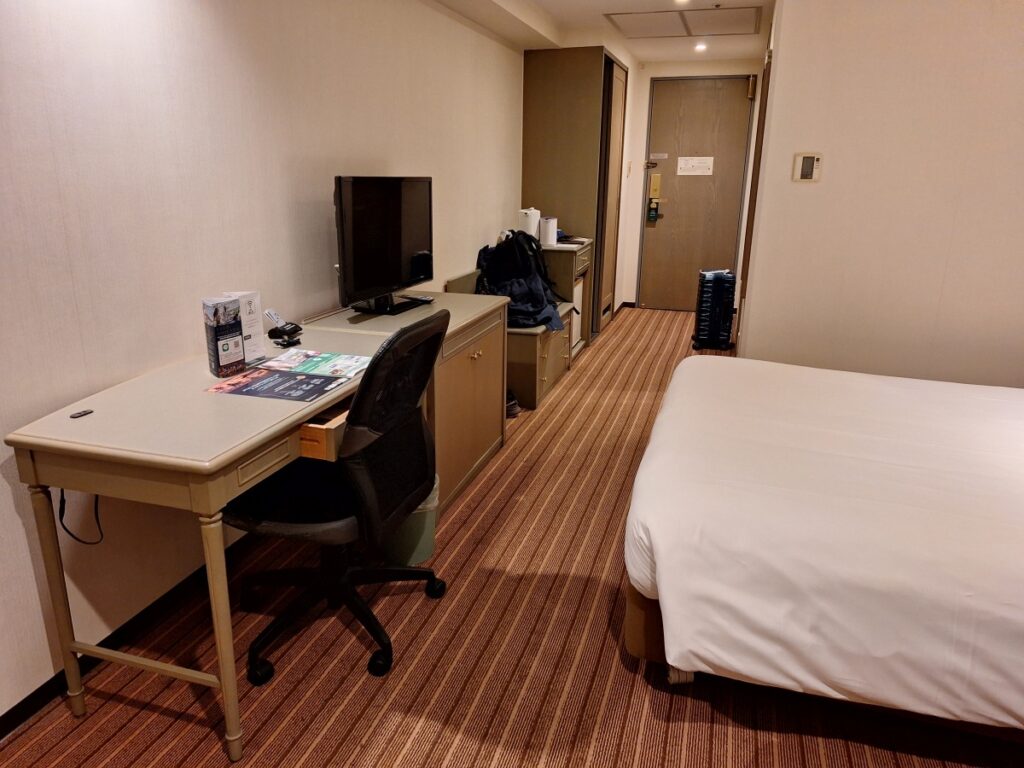 A well-lit hotel room with a double bed, large window with Japanese-style shoji blinds, a desk, and a mini fridge.