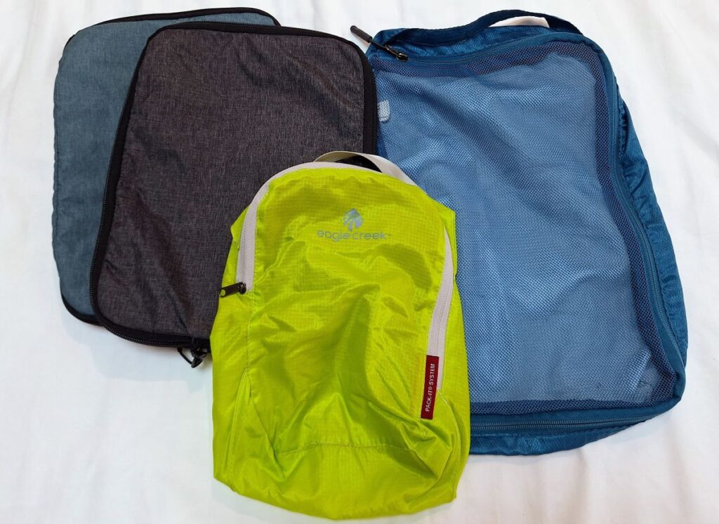 Four packing cubes of various colors. One is a Travelon soft cube in teal, one is an Eagle Creek Pack-It pouch in lime green, and two are Solo-Tourist compression cubes in black and blue.